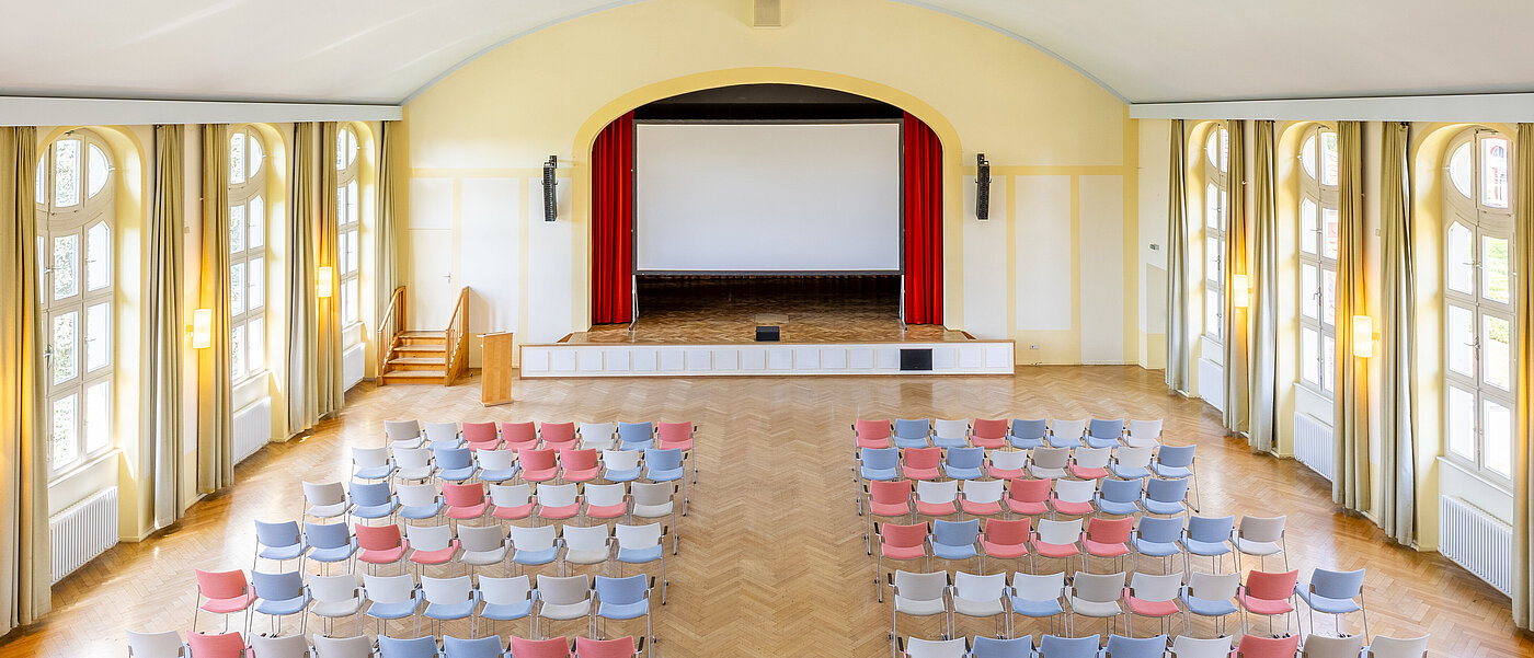 Blick von einer Empore in einen bunt bestuhlten Festsaal mit Bühne.