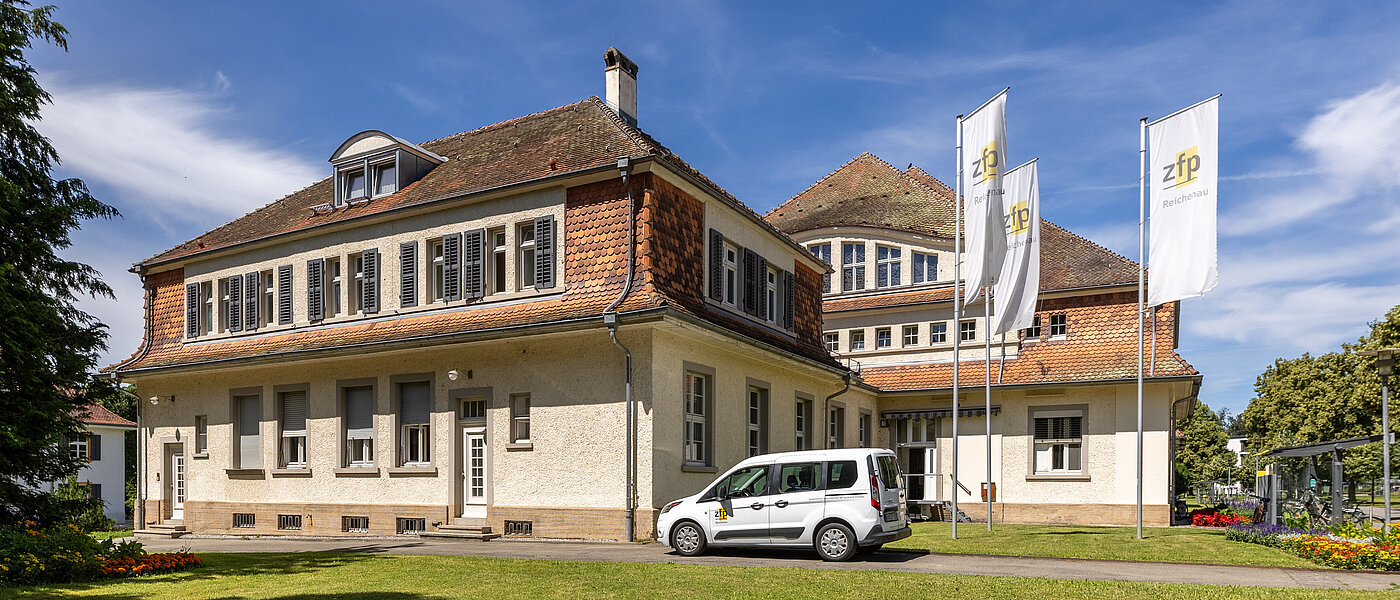 Großes Verwaltungsgebäude mit einem weißen Auto und drei weißen Fahnen davor. 